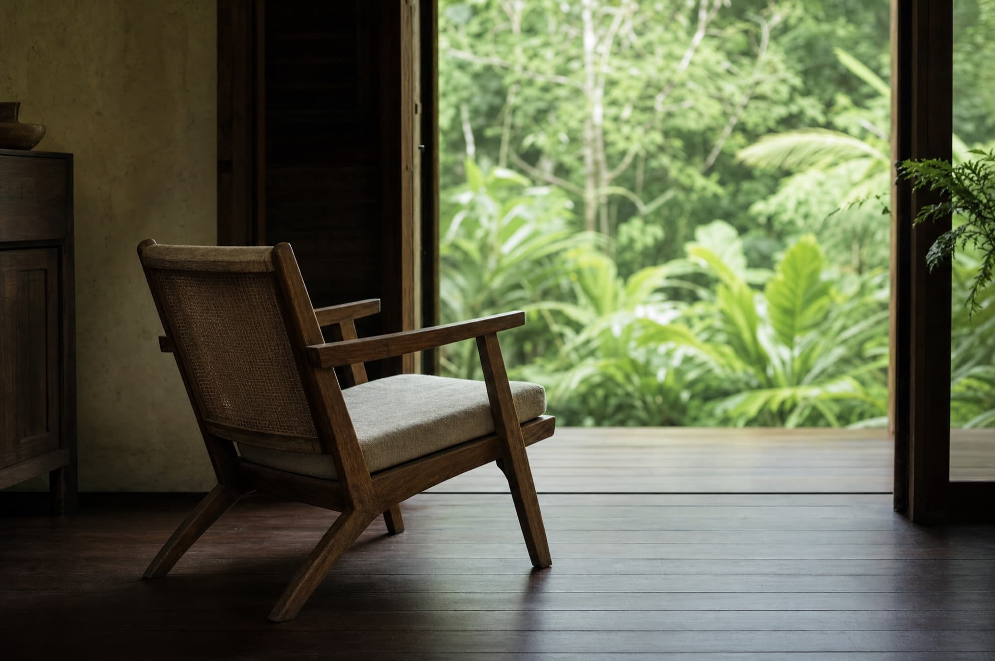 A single empty wooden chair with a linen cushion facing an open doorway onto green jungle