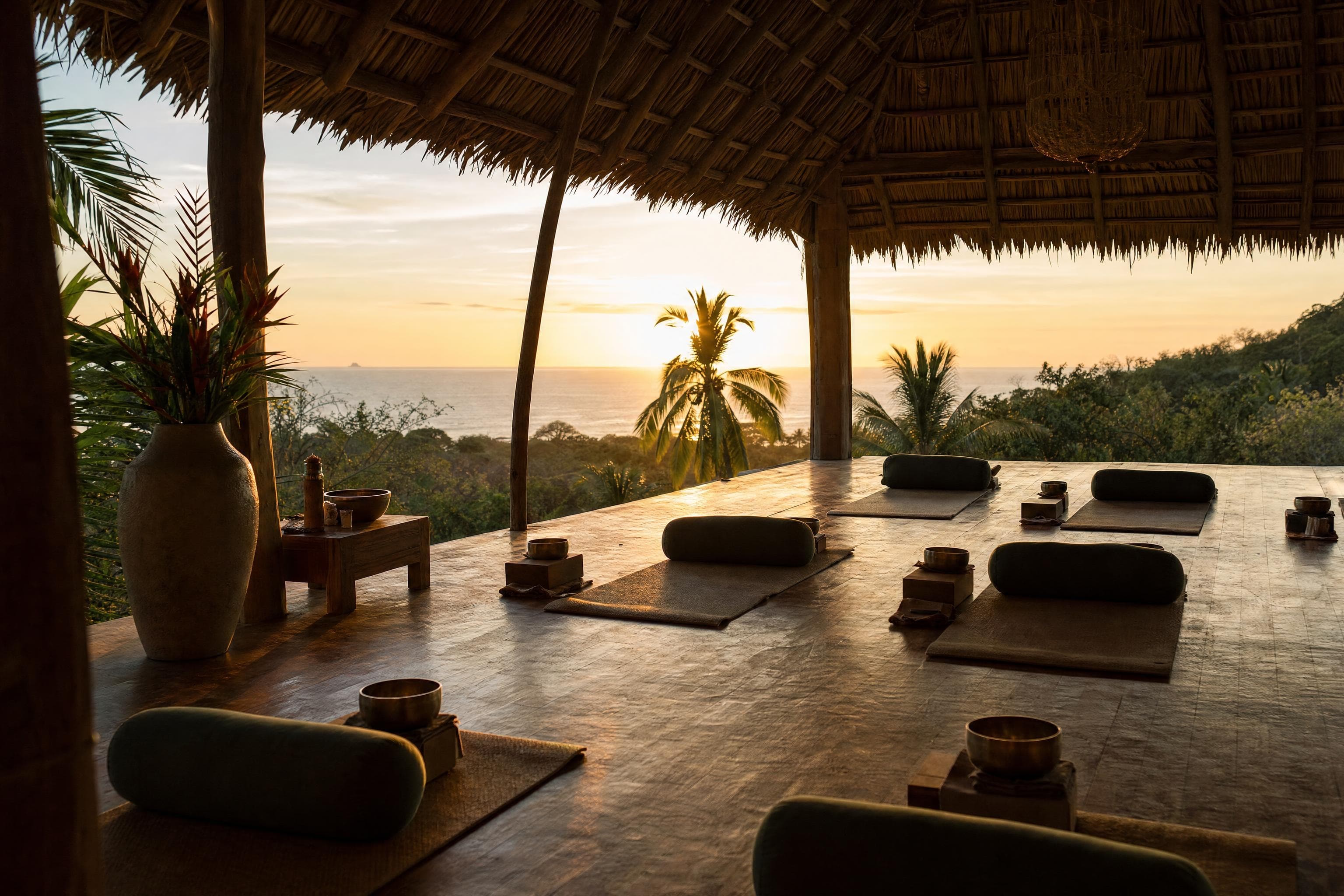 An open-air thatched yoga shala at sunset, mats and bolsters laid out toward the ocean
