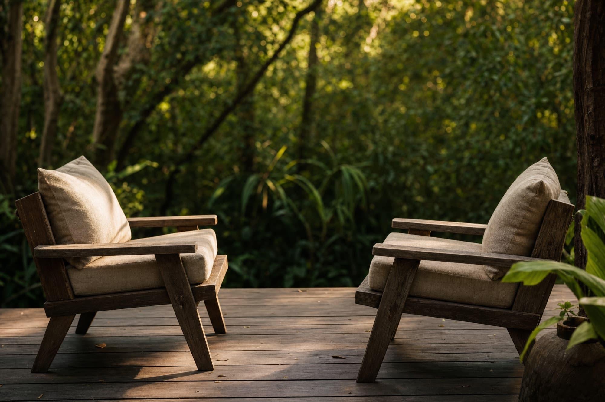 Two low wooden chairs with linen cushions facing each other on a deck at the edge of jungle, both empty
