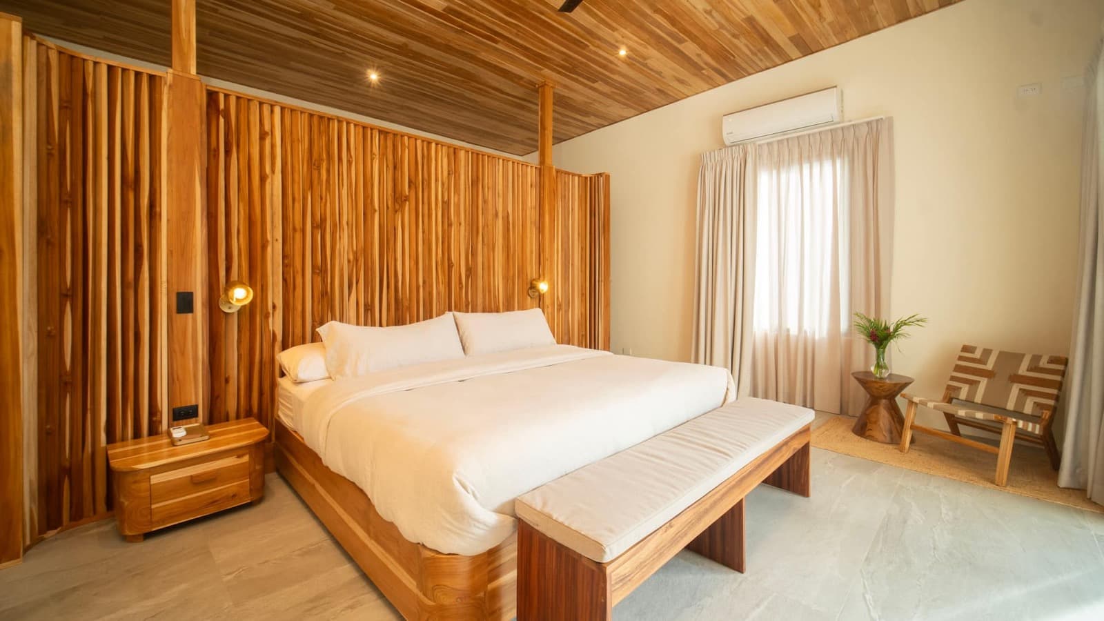 A bedroom with a king bed in white linen against a teak slat wall, brass wall sconces, and a woven lounge chair by a curtained window