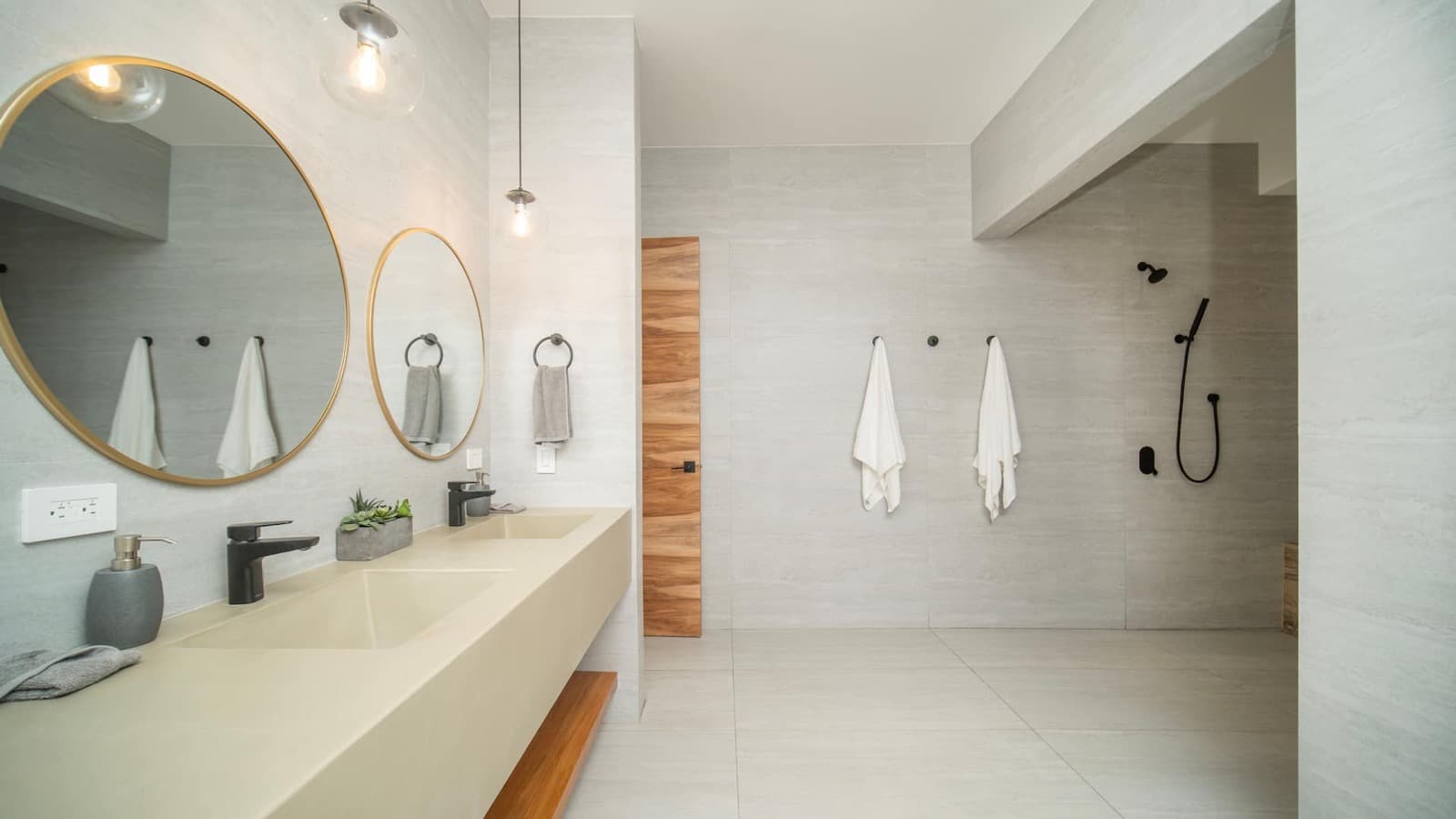 A bathroom with a long concrete double vanity, two round brass-framed mirrors, glass globe pendants, and an open walk-in shower