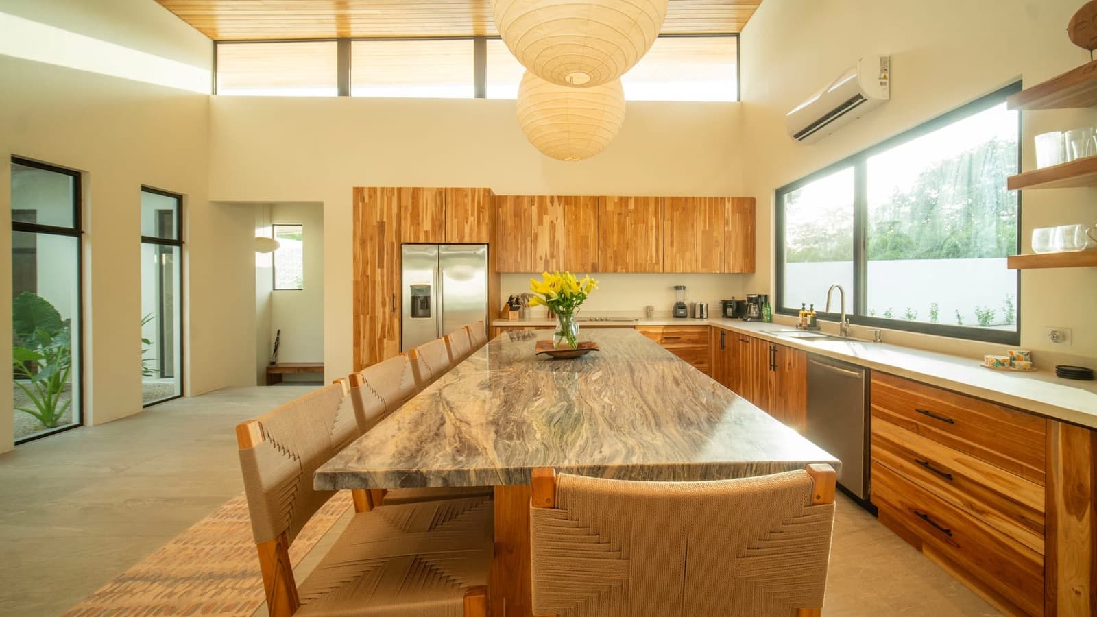 The kitchen: a marble island with six woven-back chairs under two paper lantern pendants, teak cabinetry along the window wall