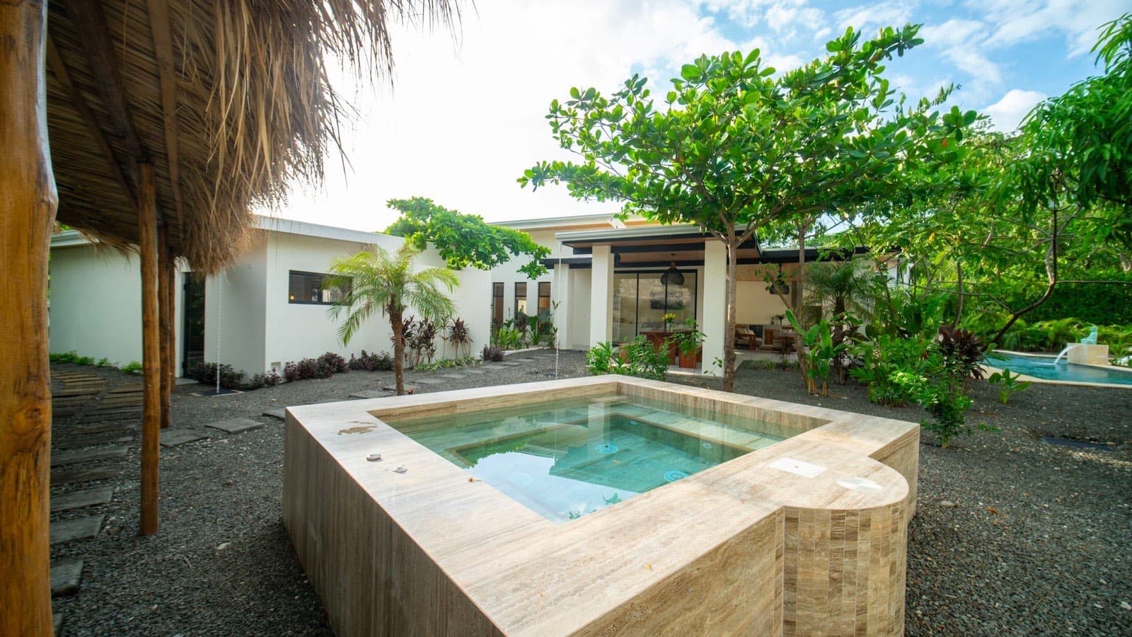 The cold plunge: a square travertine tub of clear blue water under a thatched palapa roof, mango and palm trees around it