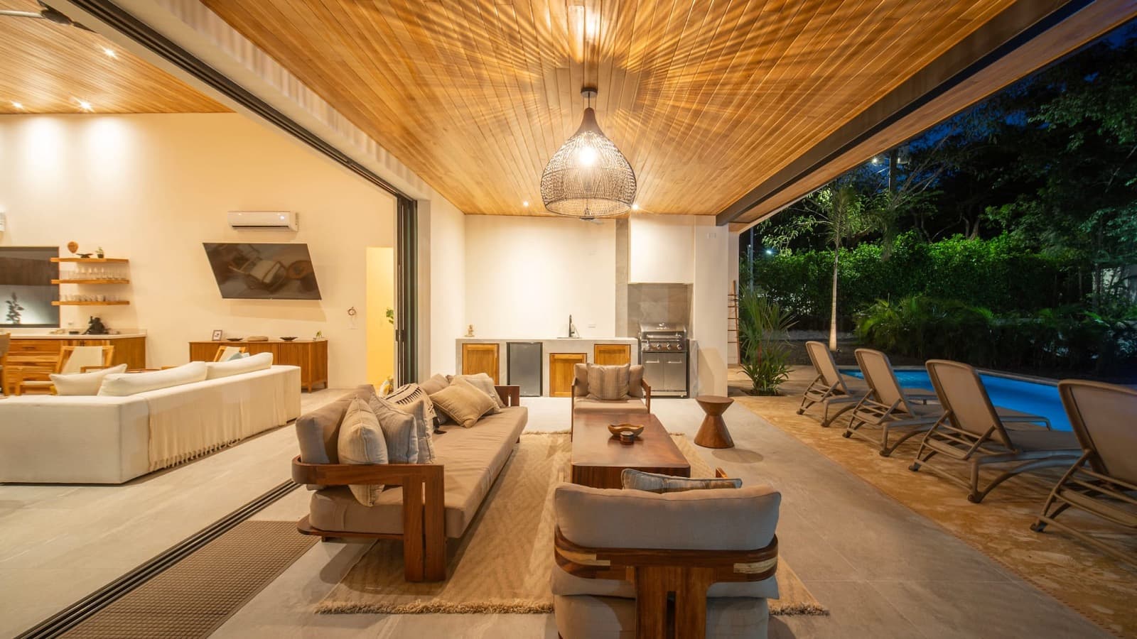 The covered open-air living room at dusk, teak-frame sofas on a jute rug, glass walls slid open to the lit pool deck on one side